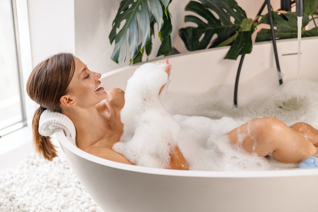 Bathing young woman relaxing in bath, smiling and playing with bubble foam